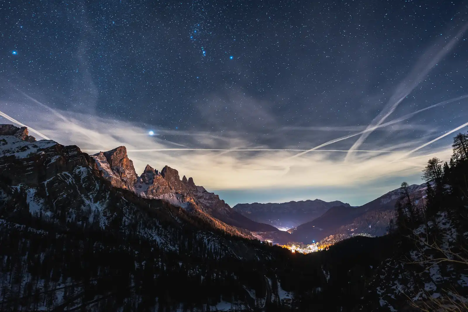 Montagne innevate sotto un cielo stellato al crepuscolo con luci di un villaggio in lontananza.