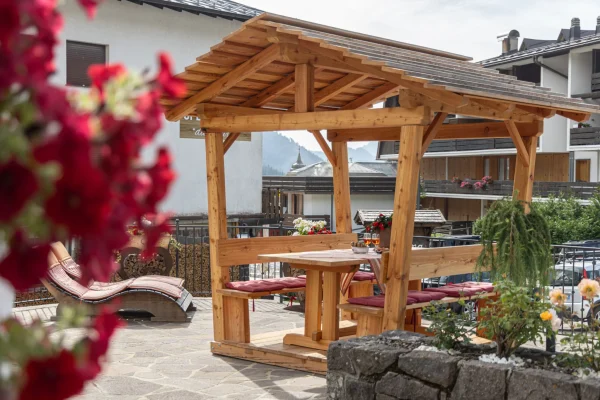 Gazebo di legno su terrazza con tavolo, sedie e fiori rossi in primo piano.