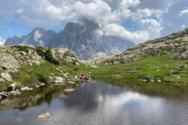 Panorama montano con laghetto riflettente, prati verdi e montagne sullo sfondo.
